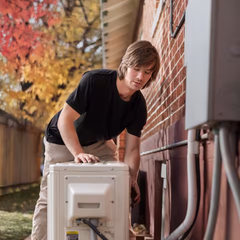 An HVAC worker fixing a heat pump next to a brick wall